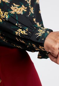 Hihan käsi mustas lillelises pluusis, millel on kollane ja roheline muster, nööbitud randme juures, kantud punase seeliku või pükste abil.