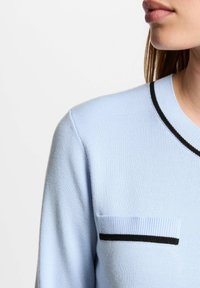 Maglione azzurro chiaro con texture a coste, dotato di colletto con bordatura nera e di una piccola tasca sul petto con un dettaglio a strisce orizzontali.