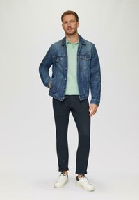 Jeansowa kurtka o klasycznym designie, w jasnoniebieskim kolorze, w połączeniu z miętową koszulką polo, ciemnymi spodniami i białymi trampkami z czarnymi akcentami.