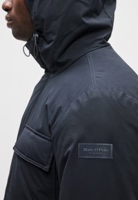 Jaqueta preta, resistente às intempéries, com capuz. Apresenta uma frente com fecho éclair, bolso no peito e um rótulo na manga. Textura suave.