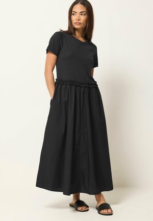 Femme debout les mains dans les poches, portant un haut noir à manches courtes et une longue jupe froncée avec des sandales noires à enfiler.