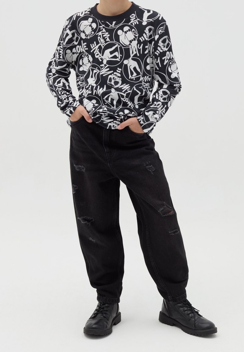 Personne portant un jean noir déchiré, des bottes noires et une chemise noire à manches longues avec des motifs graphiques blancs et du texte, debout les mains dans les poches.