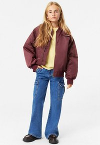 Mörkröd bomberjacka med dragkedja, ribbade muddar, kombinerad med ljusgul tröja och blå utsvängda jeans. Har sidofickor och glänsande skor.