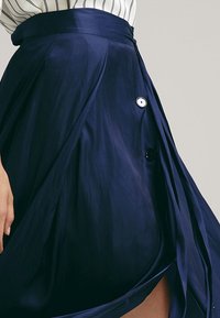 Femme portant une jupe portefeuille bleu marine avec des boutons sur une chemise blanche rayée, la main reposant près de la hanche.