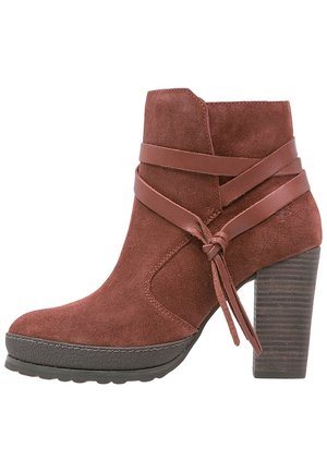 Bottine en suède couleur rouille avec un large talon empilé. Elle est dotée de lanières en cuir entourant la botte et d'un bout rond. Semelle texturée.