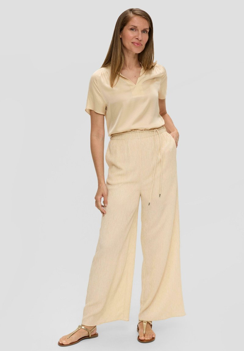 Blouse en soie beige avec un col en V et des manches courtes, associée à un pantalon large beige clair avec une taille élastique et un cordon de serrage.