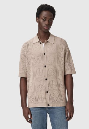 Młody mężczyzna ubrany w beżową, krótkorękawową, zapinaną na guziki koszulę dzianinową z wzorami serc i znaków pokoju, w połączeniu z niebieskimi dżinsami, stojący na szarym tle.