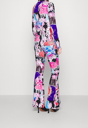 Femme portant une veste assortie vibrante et un pantalon évasé avec un imprimé tropical et voiture coloré, associé à des chaussures à talons noirs.
