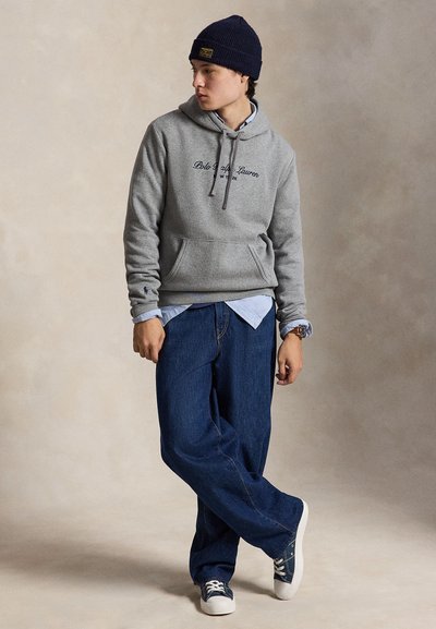 Un jeune homme porte un sweat à capuche gris, une chemise bleue, un jean ample, un bonnet en tricot et des baskets. Il se tient les jambes croisées devant un fond de studio uni.
