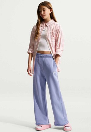 Teenager-Mädchen trägt ein weißes Crop-Top, ein pink gestreiftes Hemd, lavendelfarbene Jogginghose und pinke Slides, steht vor einem schlichten hellen Hintergrund.