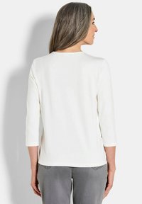 Top bianco a tre quarti con scollo rotondo, texture liscia e vestibilità rilassata, abbinato a jeans grigi su uno sfondo neutro.