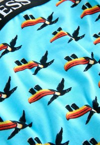 Blauer Stoff mit wiederholtem buntem Tukan-Muster und teilweise weißem Text auf schwarzem Rand.