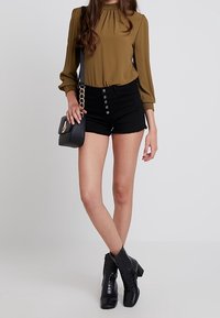 Blusa verde oliva con escote fruncido, pantalones cortos de mezclilla negros de tiro alto con cierre de botón, bolso negro y botas negras de tobillo texturizadas.