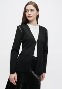 Čierny blejzer s výstrihom do V, štruktúrovaným strihom, zapínaním na jeden gombík a dvoma prednými vreckami. Ozdobený striebornými zipsami na rukávoch.