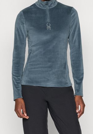 Persona que lleva un pulóver de forro polar azul grisáceo ajustado con un pequeño logo de araña en el pecho y pantalones negros.
