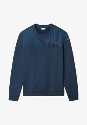 Veste matelassée noire portée sur un sweat-shirt bleu, associée à un pantalon noir texturé. Les chaussures sont noires avec des accents rouges. Tenue décontractée et superposée.