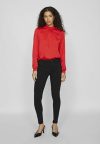 Blouse en satin rouge à manches longues avec un col haut plissé, associée à un jean noir ajusté et des escarpins noirs à bout pointu. Texture lisse sur l'ensemble.