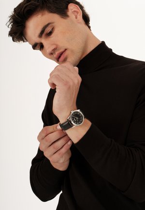 Rund ur med sort urskive, sølvdetaljer og sort læderrem. Bæreren justerer uret, mens han har en mørk striktrøje med høj hals på.