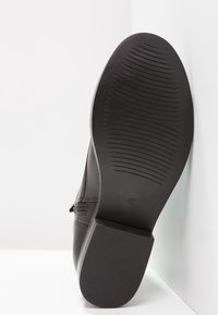 Musta kengänpohja, jossa on teksturoitu kumipohja, jossa on vaakasuoria uria pidon parantamiseksi ja koon etiketti '37' näkyvissä kannassa.