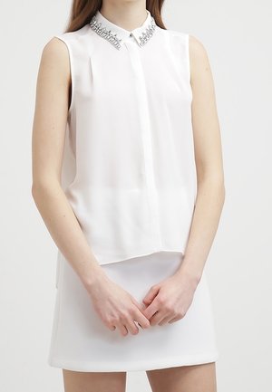 Femme portant un chemisier blanc sans manches avec un col orné et une jupe blanche, debout les mains jointes devant.
