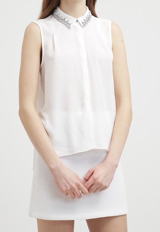 Femme portant un chemisier blanc sans manches avec un col orné et une jupe blanche, debout les mains jointes devant.