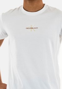 Camiseta de algodón blanca con un pequeño logotipo bordado en marrón y dorado que dice "Calvin Klein Jeans" en el pecho. Diseño de cuello redondo.