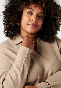 Beige gebreide trui met een kraag, V-hals ontwerp en geribbelde manchetten. De hand van het model rust op de kraag en toont gouden sieraden.