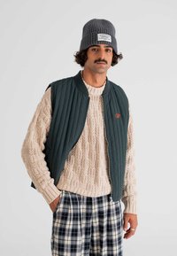 Tamnozelena kaputna prsluk s prešivanjem, preko svijetle pletive veste s kabelskim uzorkom, u kombinaciji s hlačama od kariranog platna. Model nosi sivo rebrasto kape s oznakom.