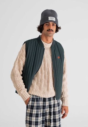 Dark green quilted vest over a cream cable-knit sweater, paired with patterned plaid trousers. Model wears a grey ribbed beanie with a label.