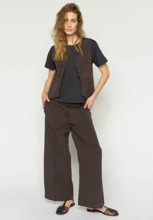 Woman with wavy hair wearing a dark t-shirt, striped vest, and wide-leg striped pants, standing with hands in pockets and black sandals.