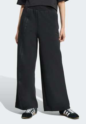 Zwarte wijde sweatpants met een soepele textuur, elastische tailleband en tonale gestreepte accenten langs de zijkanten. Gecombineerd met lage sneakers.