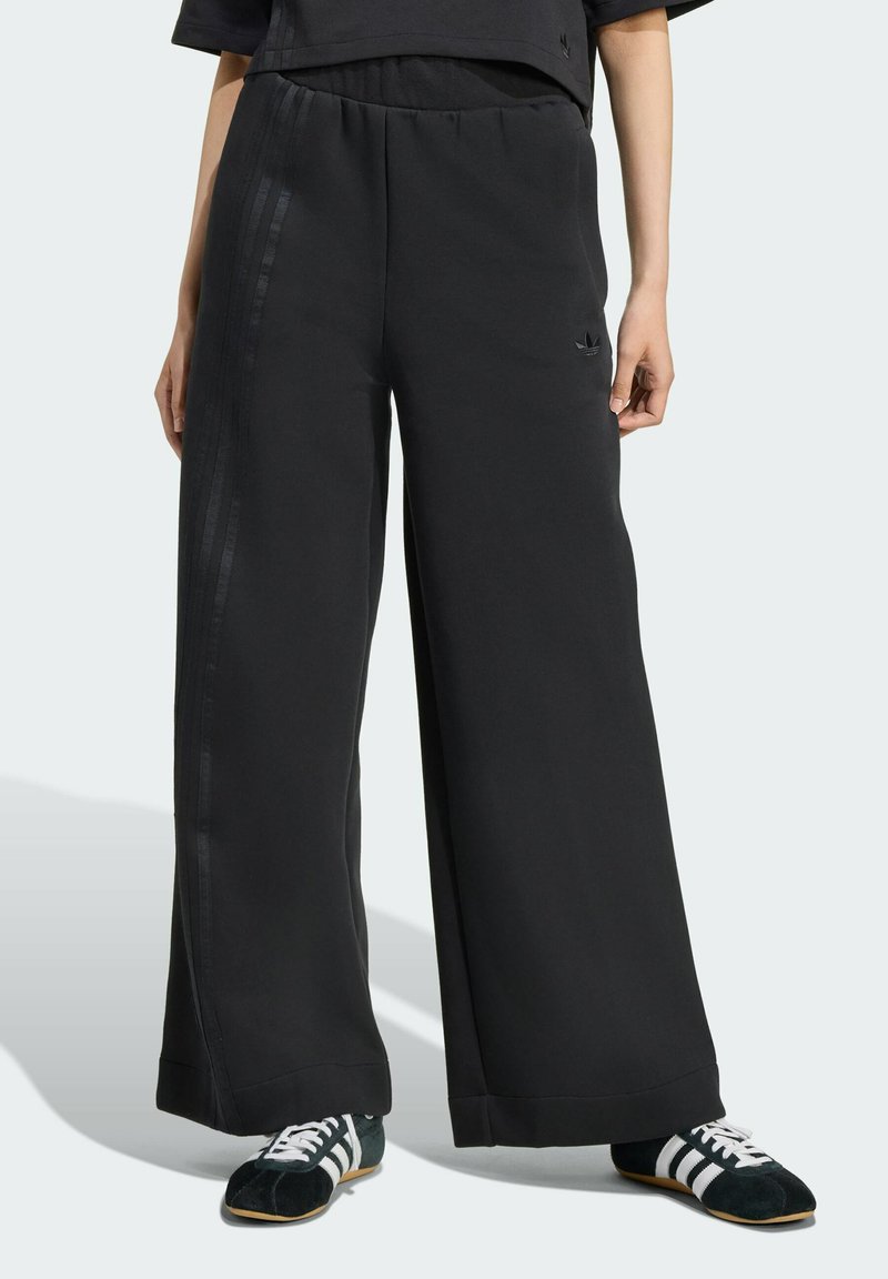 Pantalones de chándal anchos y negros con una textura suave, cintura elástica y acentos de rayas tonal a lo largo de los lados. Presentados con zapatillas bajas.