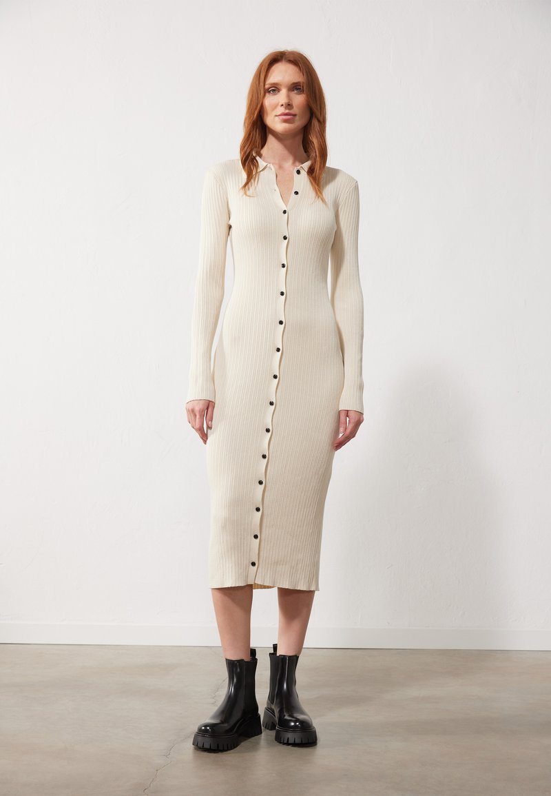 Abito in maglione a coste crema con maniche lunghe, chiusura a bottoni e spacco alto. Indossato con stivali neri con tacco spesso. Tessuto morbido e silhouette aderente.