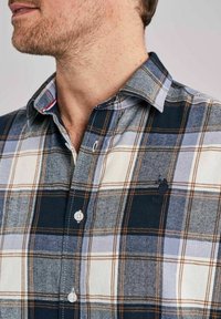 Zbliżenie mężczyzny noszącego koszulę w kratę w kolorach granatowym, kremowym i jasnoniebieskim, z starannie ułożonym kołnierzykiem i subtelnym detalem logo.