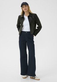 Svart läderjacka med knappar, vit t-shirt, mörkblå vida jeans och vita sneakers med svarta detaljer, återspeglades mot en vit bakgrund.