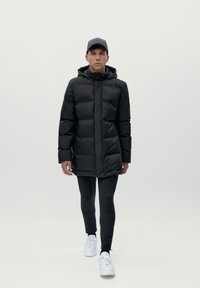 Melnā pūkaina jaka ar kapuci, nošūts dizains, priekšējais rāvējslēdzējs. Valkāta virs melnām legingām un baltām sporta apaviem, modelis stāv uz vienkārša fona.