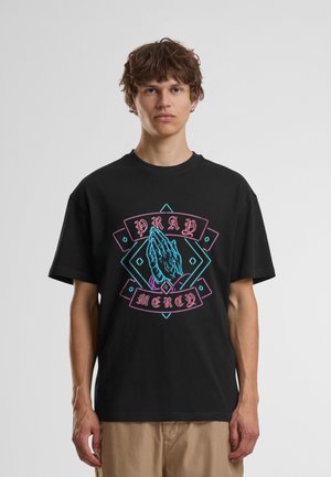Jeune homme portant un t-shirt noir avec des mains en prière style néon et les mots "Pray" et "Mercy" en design rose et bleu.