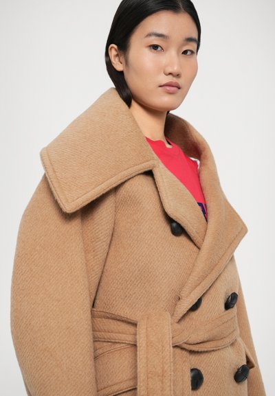 Barna gyapjúkabát nagy gallérral, dupla soros fekete gombokkal és megkötős övvel. A modell alatt vörös inget visel.