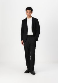 Črn blazer z dvema gumboma, bela majica s kratkimi rokavi, krojena črna hlače in črne čevlje, na enobarvni beli ozadju.