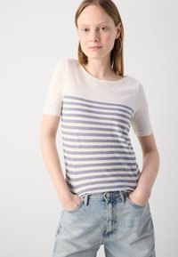 Krátky rukáv krémového trička s horizontálnymi modrými pruhmi, vyrobeného z ľahkej látky, noseného s vysokými modrými džínsami.