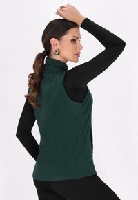 Grüne Fleece-Weste mit hohem Kragen, ärmellos und glatter Textur. Kombiniert mit einem enganliegenden schwarzen Langarmoberteil.