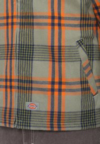 Žalias patikrintas švarkas su oranžinėmis, tamsiai mėlynomis ir geltonomis juostelėmis. Turi kišenę su pleistru ir Dickies logotipą apatinėje dalyje. Pagamintas iš švelnios medžiagos.