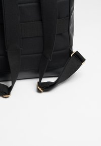 Schwarze gepolsterte Tasche aus strukturiertem Stoff, mit breiten Trägern und goldenen Hardware-Akzenten. Die Oberfläche ist glatt mit einem matten Finish.