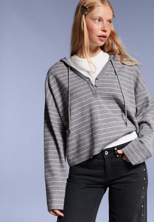 Sweat à capuche rayé gris avec un col en V et une patte de boutonnage, porté sur un haut blanc ; associé à un jean noir orné de clous.