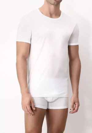 Uomo che indossa una maglietta bianca a maniche corte e boxer bianchi, in piedi contro uno sfondo bianco.