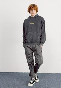 Hoodie gris surdimensionné avec un logo jaune, associé à un pantalon cargo gris. Baskets montantes noires et blanches, se tenant devant un fond uni.