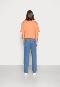 Oranžové bavlněné tričko s krátkým rukávem, v kombinaci s modrými džíny. Bílé tenisky viditelné na spodní části. Neutrální pozadí.