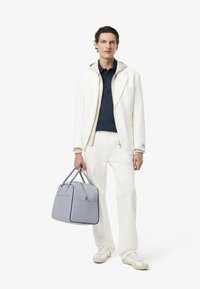 Blazer blanco sobre una polo azul marino, pantalones blancos y zapatillas crema. El sujeto sostiene un bolso de viaje gris con asas dobles.