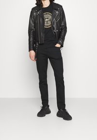 Černá kožená motorkářská bunda se stříbrným kováním, černé tričko s grafikou s kruhovým vzorem, černé džíny a černé sportovní boty.