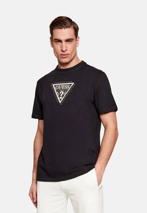 Hombre con camiseta negra de Guess con logo de triángulo y pantalones blancos, de pie con una mano en el bolsillo frente a un fondo liso.